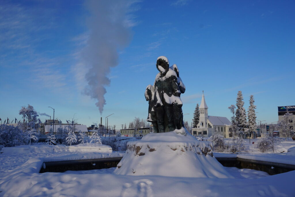 Smokestack emissions rise into the air on Feb. 7, 2025, above Golden Heart Plaza in downtown Fairbanks. Snow dusts the statute of the “Unknown First Family” that is the plaza’s centerpiece. (Photo by Yereth Rosen/Alaska Beacon)